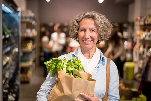 Portrait of woman looking at camera smiling carrying groceries in a paper bag while other customers are shopping at the background