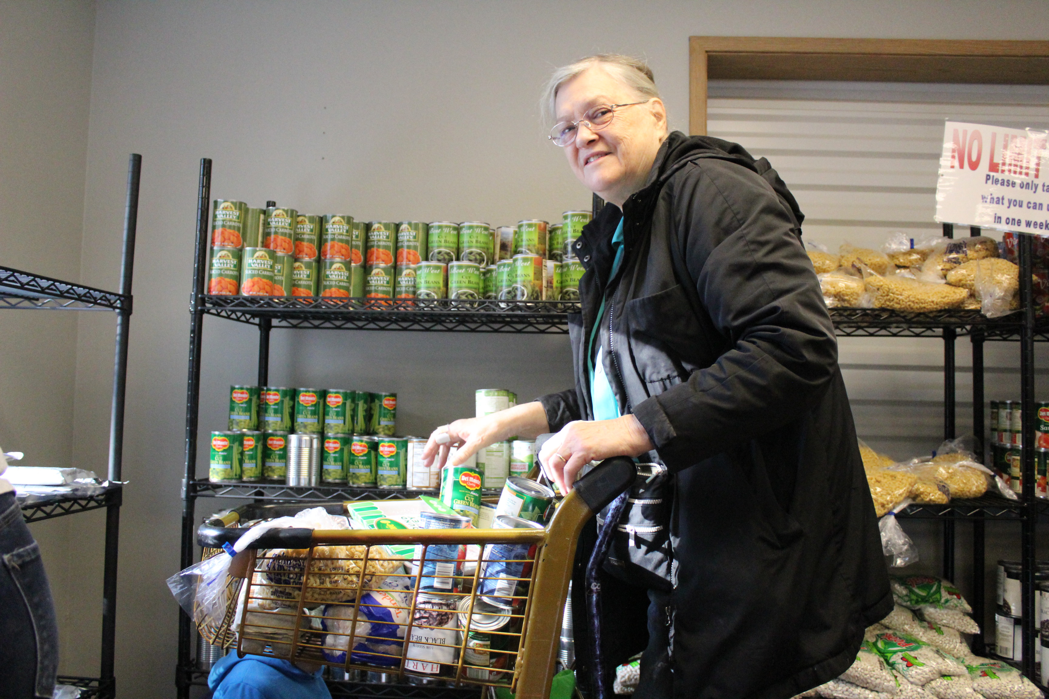 Grandma shops at local food bank. She smiles at the camera as she places canned vegetables in her cart.